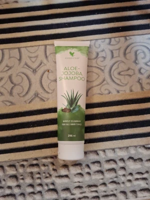 Forever Aloe-Jojoba Shampoo 296 ml - Aloe-Jojoba Shampoo från Forever i en vit och grön tub på 296 ml. Innehåller aloe vera och jojobaolja som rengör håret skonsamt. Tuben har en bild på aloe vera och text i grönt. Perfekt för dig som vill ha ett naturligt schampo med fräscha ingredienser.
