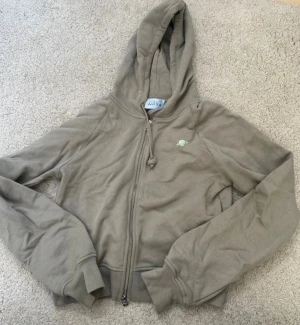 Olivgrön hoodie med planettryck - Säljer en olivgrön hoodie från PLANET NUSA  med dragkedja och huva. På bröstet finns ett litet tryck av en planet i ljusgrönt. Hoodien har långa ärmar, ribbade muddar och är gjord i mjukt bomullsmaterial. Perfekt för en chill och avslappnad stil.