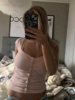 Ljusrosa topp med knappar - Säljer en ljusrosa topp med v-ringning och stora dekorativa knappar framtill. Toppen är tight och har korta ärmar, perfekt för dig som gillar en feminin och enkel stil. Passar snyggt till både jeans och kjol.