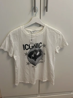 Vit t-shirt med discokula från Kappahl - Snygg vit t-shirt från Kappahl med tryck av en discokula, texten 'ICONIC' och stjärnor på framsidan. T-shirten har rund hals och korta ärmar. Perfekt för dig som gillar coola statement-plagg och vill sticka ut lite extra.