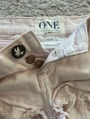 Ljusa trasiga lågmidjade jeansshorts från one teaspoon  - Säljer ett par ljusa jeansshorts från ONE by Trash Whore. Shortsen har hög midja, råa kanter och flera slitningar för en edgy look. Klassisk femficksmodell med knapp och dragkedja framtill. Perfekta för sommaren!