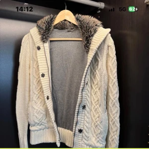 Beige stickad hoodie med fuskpäls - Stickad beige hoodie med grovt flätmönster, stora knappar och mysig huva fodrad med grå fuskpäls. Tröjan har ribbade muddar och en avslappnad passform, perfekt för kalla dagar. Inga synliga loggor eller märken.