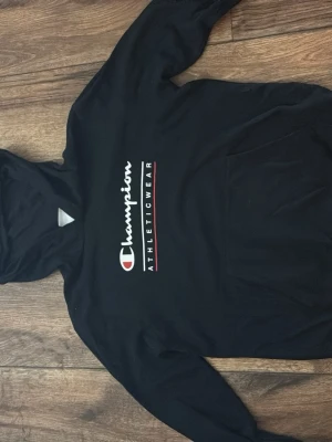 Svart Champion hoodie med logga - Säljer en svart hoodie från Champion med stor vit och röd logga på bröstet och texten ATHLETICWEAR. Hoodien har huva och en stor magficka. Perfekt för dig som gillar sportiga och avslappnade kläder.