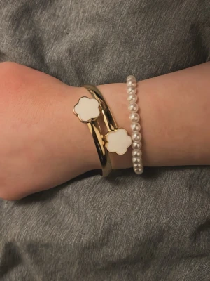 Guldigt armband med vita blommor - Snyggt armband i guld med två vita fyrklöverformade detaljer. Armbandet har en modern och stilren design som sticker ut. Perfekt för dig som gillar trendiga accessoarer med en touch av elegans.