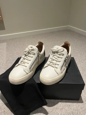 Vita sneakers från Giuseppe Zanotti - Snygga vita sneakers i läder från Giuseppe Zanotti med silverfärgade dragkedjor på sidorna och svart detalj på hälen. Klassisk låg modell med vita snören och robust sula. Perfekta för dig som gillar stilrena och exklusiva skor.