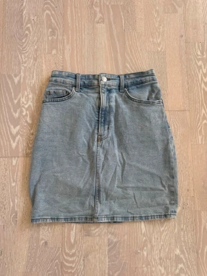 Ljusblå jeanskjol med fickor - Säljer en klassisk ljusblå jeanskjol i rak modell med fem fickor och bälteshällor. Kjolen har knapp och dragkedja framtill samt två bakfickor. Perfekt basplagg i mjukt denimtyg som passar till allt! Använd, men inga deffekter. 