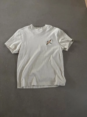 Beige t-shirt med fågeltryck - Säljer en beige t-shirt med märket Axel arigato pågrund av att jag köpte fel storlek, den är ny och aldrig använd