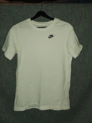 Vit Nike t-shirt med logga - Klassisk vit t-shirt från Nike med svart broderad logga på bröstet. T-shirten har rund halsringning och korta ärmar. Tillverkad i mjuk bomull för en skön och avslappnad stil. Är öppen för prisförslag vid snabb försäljning.