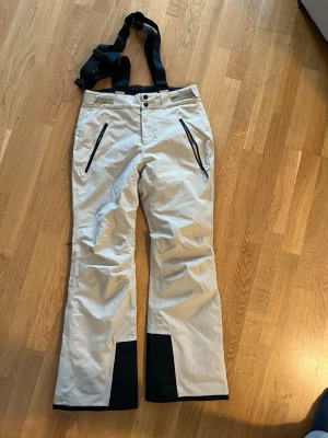 Beiga täckbyxor med svarta detaljer - Säljer ett par beiga täckbyxor med svarta förstärkningar vid bensluten och svarta dragkedjor på fickorna. Byxorna har justerbara hängslen och raka ben. Perfekta för vinterns alla äventyr i backen eller snön. Endast använda en gång.