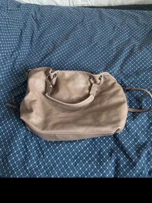 Beige axelremsväska i skinn - Snygg beige handväska i mjukt skinn med både handtag och justerbar axelrem. Väskan har en enkel, stilren design och rymlig insida. Perfekt för dig som vill ha en klassisk och praktisk väska med plats för allt du behöver. Står att den är från Å så antagligen Åhléns. Säljer även den på vinted 