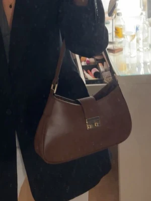 Brun handväska  - Snygg brun handväska med axelrem och guldfärgat spänne framtill. Väskan har ingen dragkedja utan stängs med spännet bara 💞