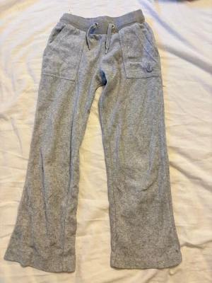 Gråa mjukisbyxor Juicy Couture - Mysiga gråa mjukisbyxor från Juicy Couture i mjuk velour. Byxorna har flare brn, bred resår i midjan med dragsko och två stora fickor fram. Klassisk Juicy Couture-logga broderad på ena fickan. Det är barn storlek, 5-6år men är väldigt små i storleken. Knapparna där bak har tappat färgen lite men inget som syns så jättemcyket direkt.