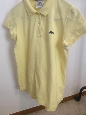 Gul pikétopp från Lacoste - Klassisk gul pikétopp från Lacoste med korta ärmar och krage. Toppen har knappar framtill och den ikoniska gröna krokodilloggan på bröstet. Tillverkad i bomull för en bekväm och luftig känsla.