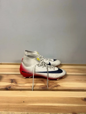 Nike Mercurial vita fotbollsskor - Säljer ett par Nike Mercurial fotbollsskor med högt skaft. Skorna är vita med röda och mörkblå detaljer samt gula inslag vid snörningen. Tillverkade i syntetmaterial för bra grepp och kontroll på planen. Perfekta för dig som vill ha stilrena och snabba skor på matchen.