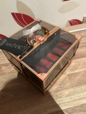 Technic sminkväska med läppstift - Snygg sminkväska från Technic i svart och roséguld med transparent lock och handtag. Innehåller flera röda läppstift. Perfekt för att organisera och förvara sminkprodukter på ett stilrent sätt.