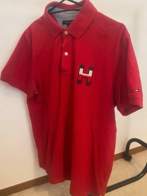 Röd pikétröja från Tommy Hilfiger - Klassisk röd pikétröja från Tommy Hilfiger med korta ärmar och krage. Tröjan har två knappar framtill och en stor H-logga på bröstet. Tillverkad i mjuk bomull som känns skön mot huden. Perfekt för en avslappnad och stilren look.