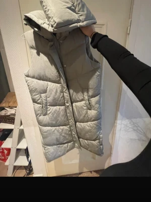 Ljusgrå lång dunväst med huva - Säljer en ljusgrå, quiltad dunväst med avtagbar huva och dragkedja framtill. Västen har två stora fickor på sidorna och en längre, oversized passform som ger en riktigt trendig look. Perfekt att slänga över en hoodie för extra värme.