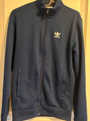 Mörkblå zip hoodie från Adidas - Snygg mörkblå hoodie från Adidas med dragkedja framtill och två fickor. Klassisk Adidas-logga i vitt på bröstet. Tillverkad i mjukt material som känns skönt mot huden. Perfekt för en avslappnad streetwear-look.