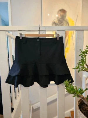 Svart volangkjol från Zoul - Snygg svart kortkjol från Zoul med dubbla volanglager nedtill. Kjolen har bälteshällor, knapp och dragkedja framtill. Perfekt för dig som vill ha en enkel men trendig kjol med volangdetalj.