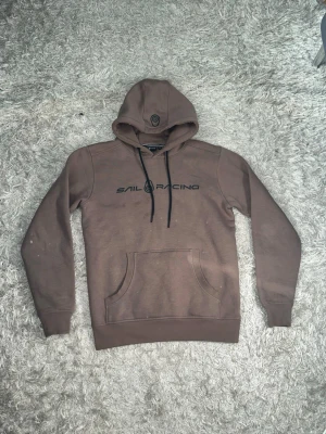 Brun hoodie från Sail Racing - Snygg brun hoodie från Sail Racing med svart logga på bröstet och klassisk känguruficka. Huva med svarta dragsnören och ribbade muddar vid ärmar och nederkant. Perfekt för en avslappnad och sportig stil. Varan är i bra skick, för fler bilder/frågor skriv privat, storlek S/M