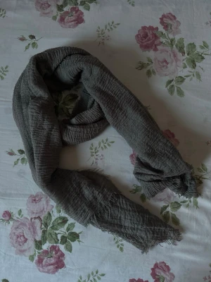 Grå crinklad halsduk  - En stilren grå halsduk med crinklad struktur och fransiga kanter. Perfekt att svepa runt halsen för en avslappnad look.