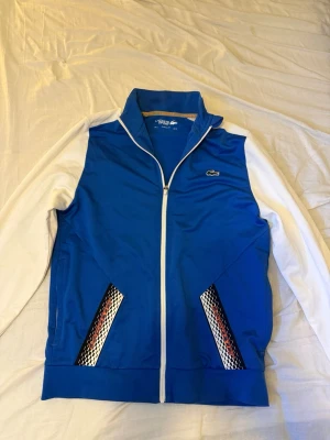 Blå och vit ziptröja från Lacoste - Snygg blå ziptröja från Lacoste med vita ärmar och vita detaljer. Köptes för runt 2 tusen, använt ett fåtal gånger. Storlek M men passar även S (lite liten i storlek) Priset går att diskutera. 