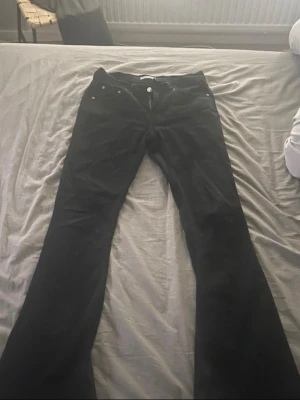 Svarta bootcut jeans - Säljer ett par svarta bootcut jeans med klassisk femficksdesign och knappgylf. Jeansen har vida ben och detaljer i metall vid fickorna. Perfekta för dig som gillar en retroinspirerad look.