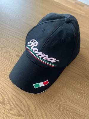 Svart keps med Roma-brodyr - Snygg svart keps med broderad text 'Roma' i rosa och italienska flaggans färger på skärmen. Kepsen har även en liten italiensk flagga broderad framtill. Justerbar baktill 💕