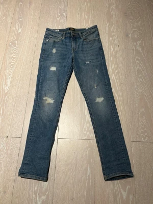 Slim jeans från Jack & Jones - Säljer ett par blå jeans från Jack & Jones med skinny fit och slitna detaljer på benen. Jeansen har klassisk femficksmodell, normal midja och är tillverkade i bomull med lite stretch för extra komfort. Perfekta för en avslappnad och trendig look.