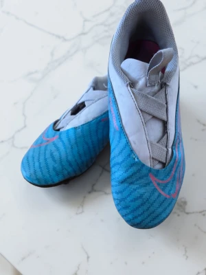 Blå Nike fotbollsskor med rosa detaljer - Säljer ett par blå Nike fotbollsskor med rosa swoosh och mönstrad ovandel. Skorna har en modern, strömlinjeformad design med elastisk snörning och grå detaljer. Perfekta för dig som vill sticka ut på planen och ha bra grepp. Tillverkade i syntetmaterial.