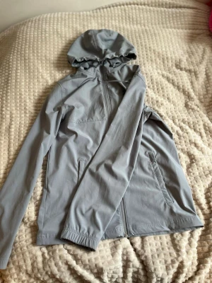 Ljusgrå vindjacka från Under Armour - Snygg ljusgrå vindjacka från Under Armour med huva och dragkedja framtill. Jackan har två sidofickor med dragkedja och en diskret logga på bröstet. Tillverkad i ett lätt och vindtätt material som passar perfekt för blåsiga dagar.