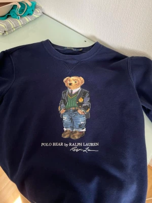 Mörkblå Polo Bear sweatshirt Ralph Lauren - Säljer en mörkblå sweatshirt från Ralph Lauren med den ikoniska Polo Bear-trycket på framsidan. Tröjan har rund halsringning och är tillverkad i mjukt bomullsmaterial. Perfekt för dig som gillar klassisk och stilren streetwear med en twist.