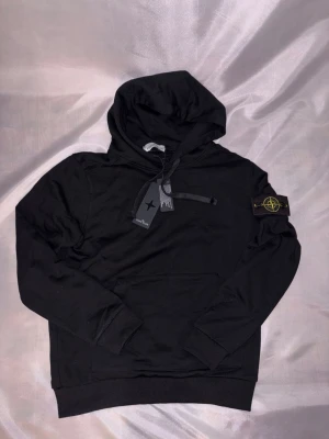 Svart hoodie från Stone Island - Snygg svart hoodie från Stone Island med klassisk patch på vänster ärm. Hoodien har huva med dragsko och en stor magficka framtill. Tillverkad i mjukt bomullsmaterial som känns skönt mot huden. Perfekt för en avslappnad streetwear-look.