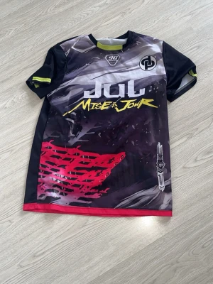 Svart och röd Jul t-shirt - Unik t-shirt från Jul med coolt grafiskt tryck i svart, rött, gult och grått. Framsidan har texten 'Miser JouR' och baksidan har logga och texten 'Nineta'. T-shirten har korta ärmar och är tillverkad i ett syntetmaterial som känns sportigt och lätt.