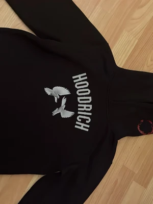 Svart HOODRICH hoodie med duvor - Säljer en svart hoodie från HOODRICH med tryck av vita duvor och texten 'HOODRICH' på framsidan. På ryggen finns ett stort tryck med fler duvor och texten 'From Nothing To Something' i rött samt 'PRODUCT OF THE STREETS EST. 2014' nedtill. Klassisk huva och mjukt material. 