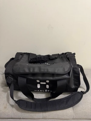 Svart duffelbag från Haglöfs Lava30 - Säljer en svart duffelbag från Haglöfs med stort huvudfack, justerbar axelrem och bärhandtag. Väskan har vit logga framtill och är tillverkad i slitstarkt material, perfekt för resor eller gymmet. Snygg och praktisk design med flera fack för enkel organisering.
