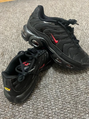 Nike Air Max Plus TN  Strl:41 - Säljer ett par svarta Nike Air Max Plus sneakers med röda detaljer och TN-logga på hälen. Skorna har en mesh- och syntetöverdel, synliga Air-enheter i sulan och en sportig design med vågiga linjer. Perfekta för dig som gillar streetwear och vill sticka ut.