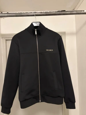 Svart jacka med dragkedja Les Deux - Snygg svart ziptröja från Les Deux med hög krage och diskret logga på bröstet. Tröjan har hel dragkedja, två fickor framtill och ribbade muddar vid ärmslut och nederkant. Perfekt för en clean och stilren look. Nypris 1299kr. Kan gå ner i pris vid en snabb affär!