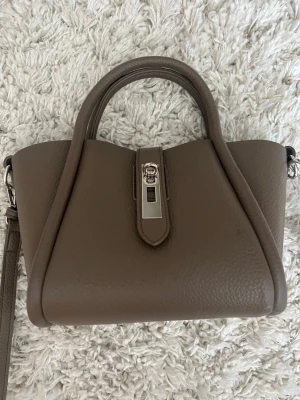 Gina Tricot brun/beige handväska - Snygg handväska i mjukt skinn med silverfärgat spänne framtill. Väskan har både handtag och justerbar axelrem, samt en stilren och modern form. Perfekt för dig som gillar minimalistisk design och neutrala färger.