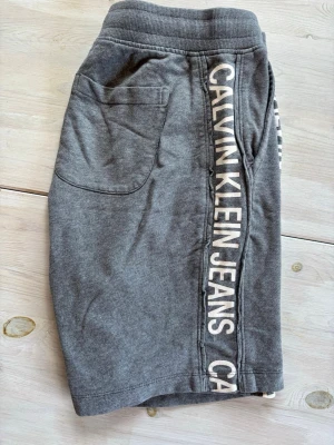 Grå shorts från Calvin Klein Jeans - Snygga grå shorts från Calvin Klein Jeans med vit logga längs sidorna. Modellen har elastisk midja och två fickor fram. Perfekta för en avslappnad och trendig stil under varmare dagar.
