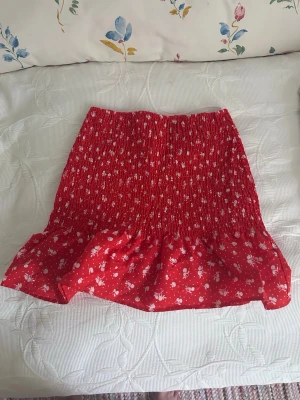 Röd blommig kortkjol med volang från HM - Söt röd kortkjol med vita små blommor och volangkant. Kjolen har smock i midjan för en bekväm passform och är perfekt för varma dagar. Stretchig så att den kan passa flera storlekar, inte bara XS utan även S. Materialet känns lätt och luftigt. Från divided HM.