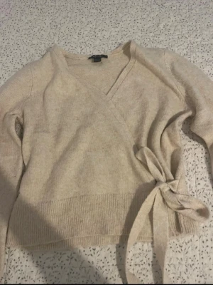 Beige omlottstickad tröja - Snygg beige stickad omlott-tröja med knytband i midjan och långa ärmar. Tröjan har ribbstickade muddar och en mjuk, mysig känsla. Perfekt för lager-på-lager och enkel att styla till olika outfits.