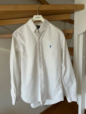 Ralph Lauren skjorta - Klassisk vit skjorta med button-down krage och lång ärm. Skjortan har en liten blå broderad logga på bröstet och stängs med vita knappar framtill. Tillverkad i bomull och har en tidlös, rak passform.      Priset är inte skrivet i sten
