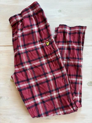 Rödrutiga pyjamasbyxor Lyle & Scott - Mysiga pyjamasbyxor från Lyle & Scott i röd, marinblå och vit rutigt mönster. Byxorna har resår i midjan med dragsko och en liten gul logga på benet. Perfekta för chillkvällar hemma.