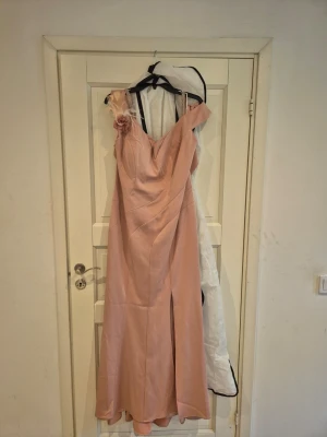 Lång ljusrosa axelbandslös klänning - Elegant långklänning i ljusrosa med offshoulder-design och en dekorativ blomdetalj på ena axeln. Klänningen har en slits framtill och är tillverkad i ett glansigt material som ger en lyxig känsla. Perfekt för speciella tillfällen. Storlek 44-46