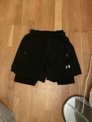 Svarta träningsshorts Under Armour - Säljer ett par svarta träningsshorts från Under Armour med snörning i midjan och dragkedjefickor. Shortsen har en diskret logga på ena benet och innerbyxa för extra komfort. Perfekta för gymmet eller löprundan.