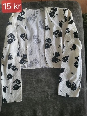Vit blommig kofta från Eurofashion - Kort, öppen kofta i vit strukturvävd textil med svarta blommor. Långa ärmar och lätt boxig passform. Perfekt för att lyfta en enkel outfit med ett coolt mönster.