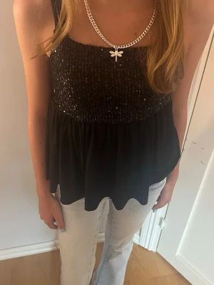 Svart glittrig festtopp - Snygg svart topp med glittriga detaljer upptill och smockad byst. Toppen är ärmlös och har ett luftigt fall nedtill, perfekt för en trendig look. Passar till både jeans och kjol för en cool outfit.