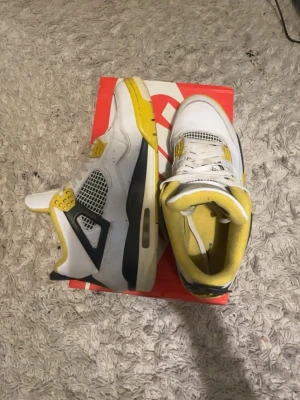 Nike Air Jordan 4 Retro gul/vit - Nike Air Jordan 4 Retro sneakers i vitt och gult med svarta detaljer. Skorna har meshpaneler på sidorna, synlig Air-sula och klassisk Jumpman-logga på hälen. Ovandelen är i läder och mesh, och innersulan är gul vadderad textil.