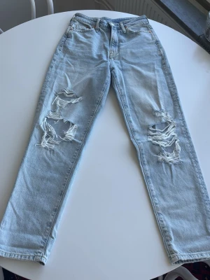 Ljusblå ripped mom jeans - Säljer ett par ljusblå mom jeans med raka ben och stora slitningar på båda knäna. Jeansen har klassisk femficksmodell och hög midja. Tillverkade i denim med en avslappnad passform, perfekta för en trendig och avslappnad stil.